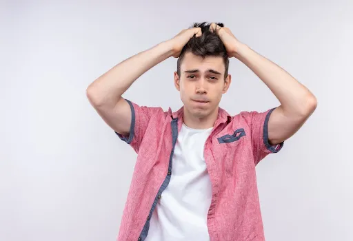 Teenage boy in a pink shirt looking stressed while pulling his hair with both hands against a plain light background.