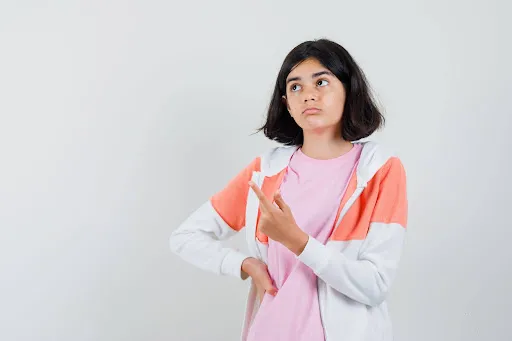 “Thoughtful teenage girl in casual clothes looking upward with a confused expression while gesturing with her hands against a plain gray background.