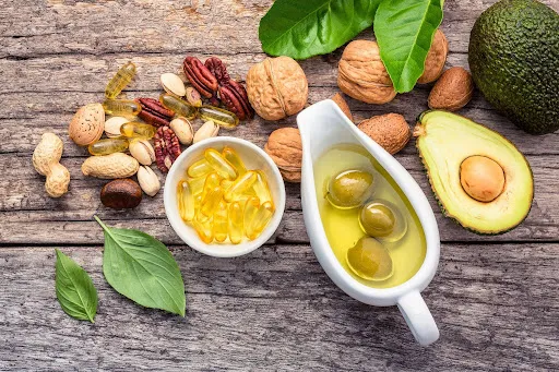 Healthy fats and nutrition supplements including avocado, olive oil, omega-3 capsules, nuts, and seeds arranged on a wooden surface, representing hair and wellness nutrition